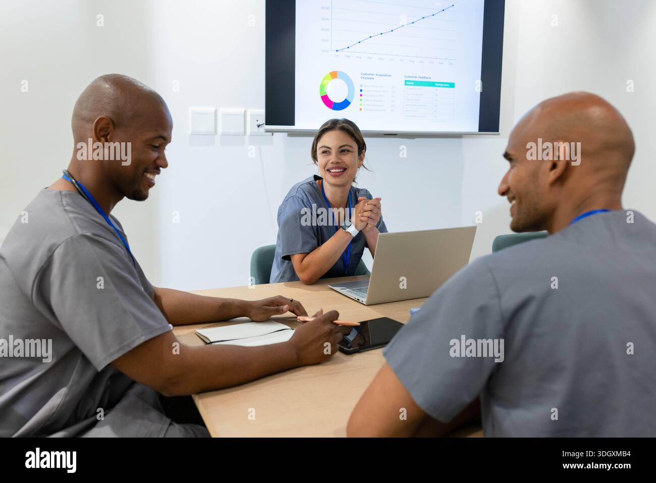 Afroamerikanisches Ärzteteam in grauen Scrubs sitzt in einem Meeting, eine Frau präsentiert sich auf einem Laptop-Monitor Stockfoto