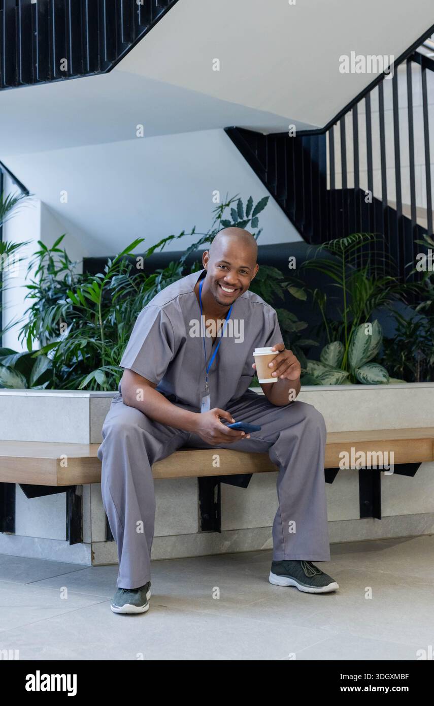 Erwachsener Mann, der auf einer Vorhof-Bank sitzt und graue Schrubben trägt und Kaffee und Telefon mit blauem Schlüsselband hält Stockfoto