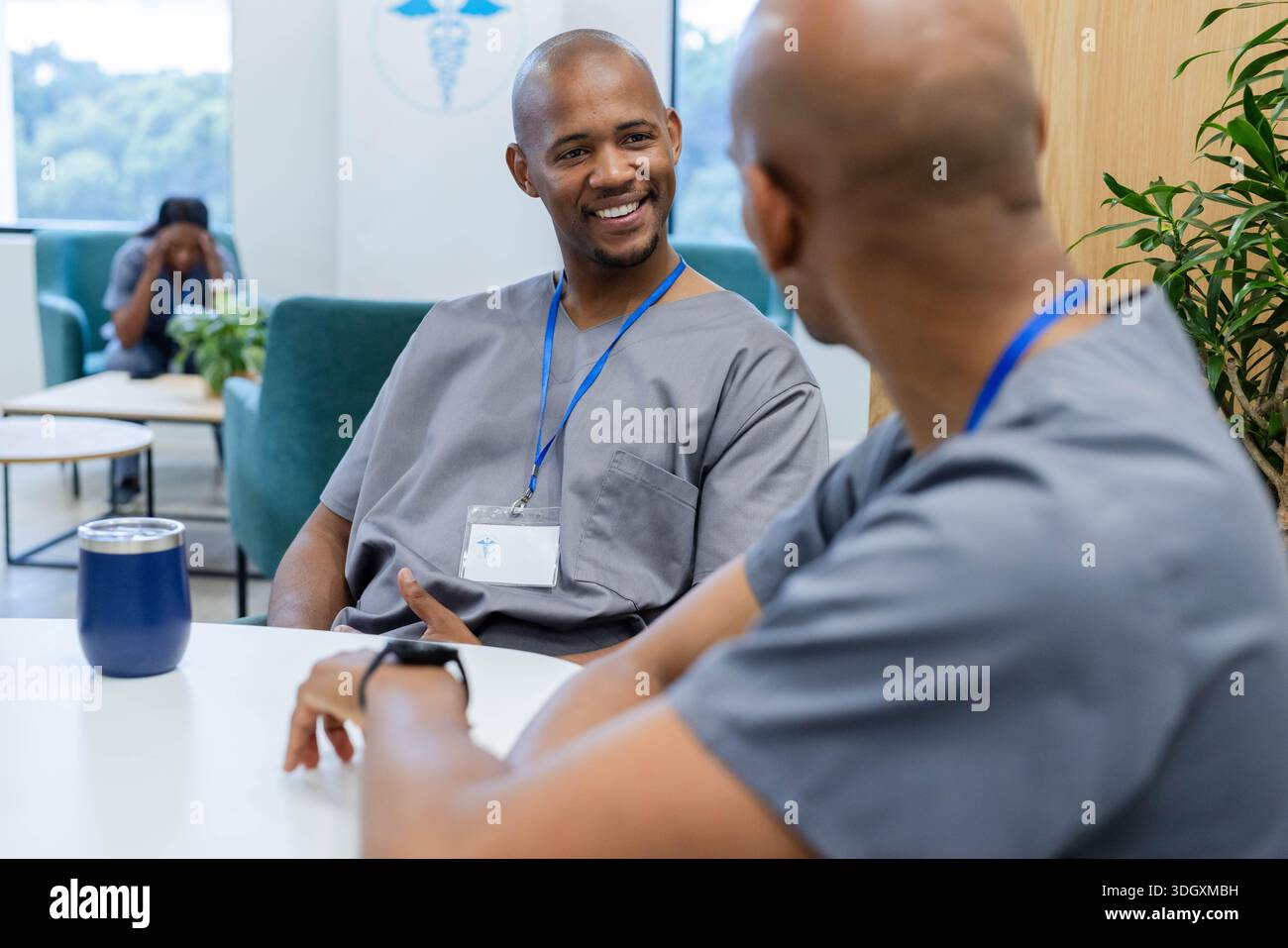 Afroamerikanische männliche Kollegen sitzen in der Klinik-Lounge, in grauen Schutten mit blauem Tumbler Stockfoto