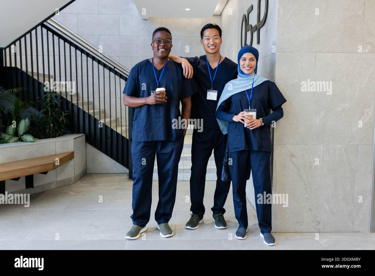 Verschiedene Kollegen lächeln, halten Kaffeetassen, tragen marineblaue Peelings und blaue Schlüsselbänder in der Klinik Stockfoto