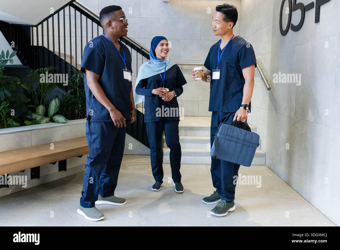 Verschiedene Gesundheitsfachkräfte in der Marine schrubben stehen und plaudern in der Lobby der Klinik, halten eine Kaffeetasse Stockfoto