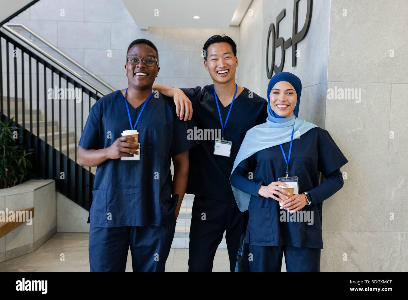 Verschiedene medizinische Mitarbeiter in marineblauen Schutten stehen in der Lobby des Krankenhauses mit Bechern und blauen Ausweisen Stockfoto
