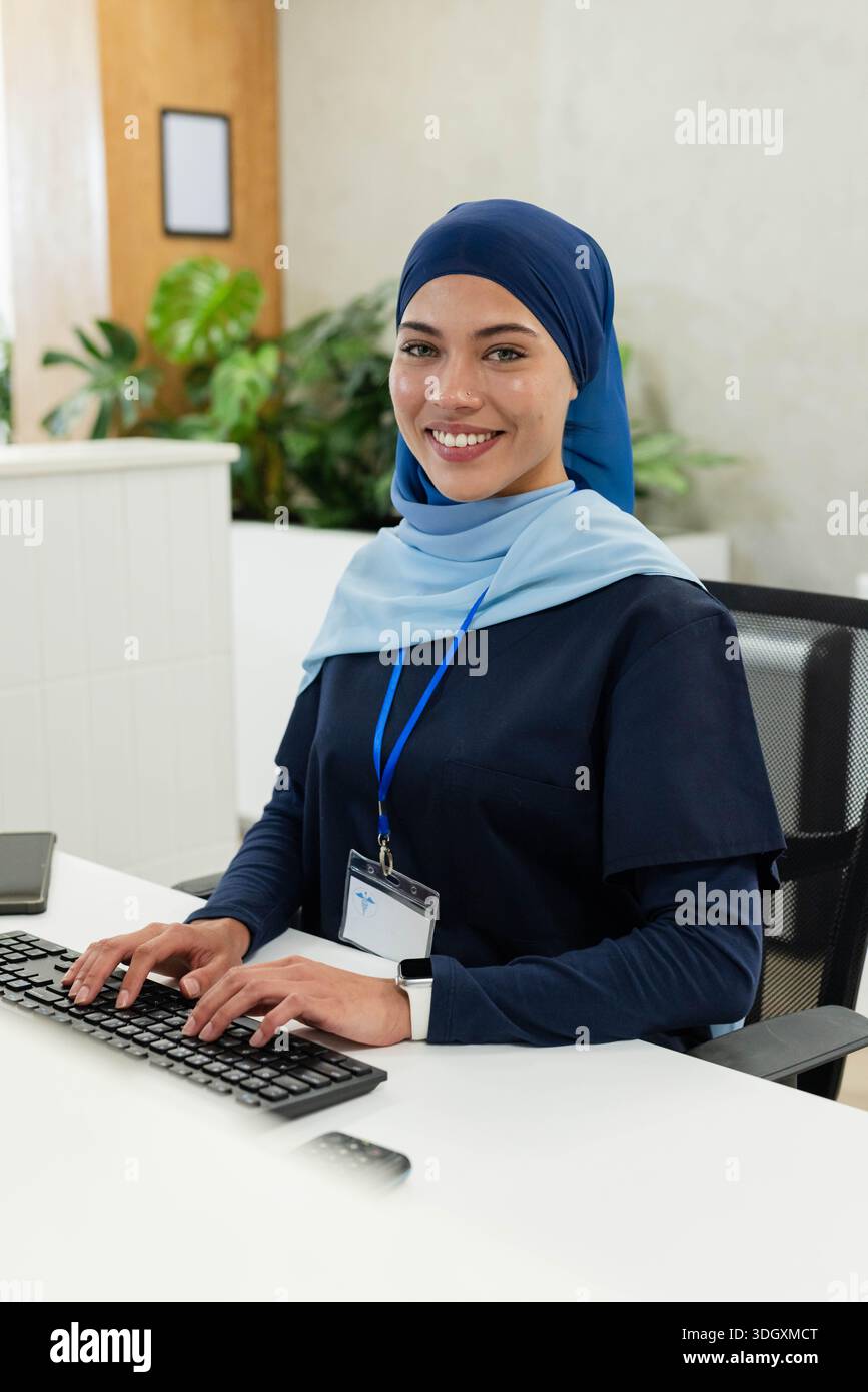 Afroamerikanische Frau, die am Rezeptionstresen auf der Tastatur in marineblauen Scrubs und blauem Lanyard-Abzeichen tippt Stockfoto