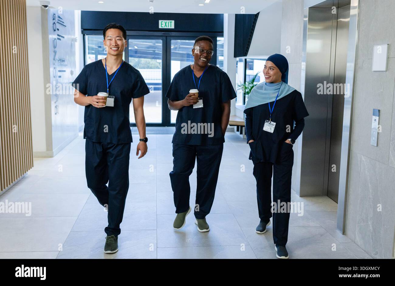 Verschiedene Kollegen, die den Krankenhausflur hinunterlaufen und To-go-Kaffeetassen halten und marineblaue Peelings tragen Stockfoto