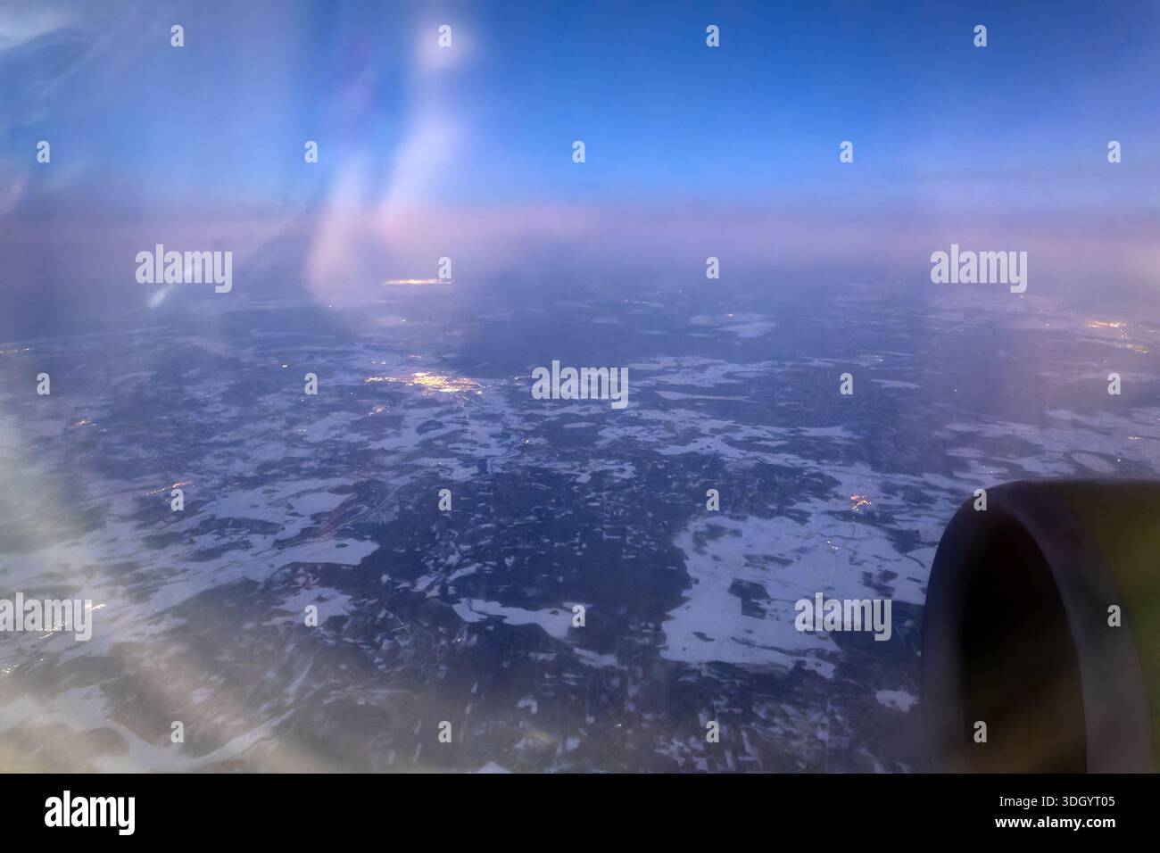 Luftaufnahme aus dem Flugzeugfenster während der Winterdämmerung. Schneebedeckte Landschaft, Stadtlichter und Motor sichtbar Stockfoto