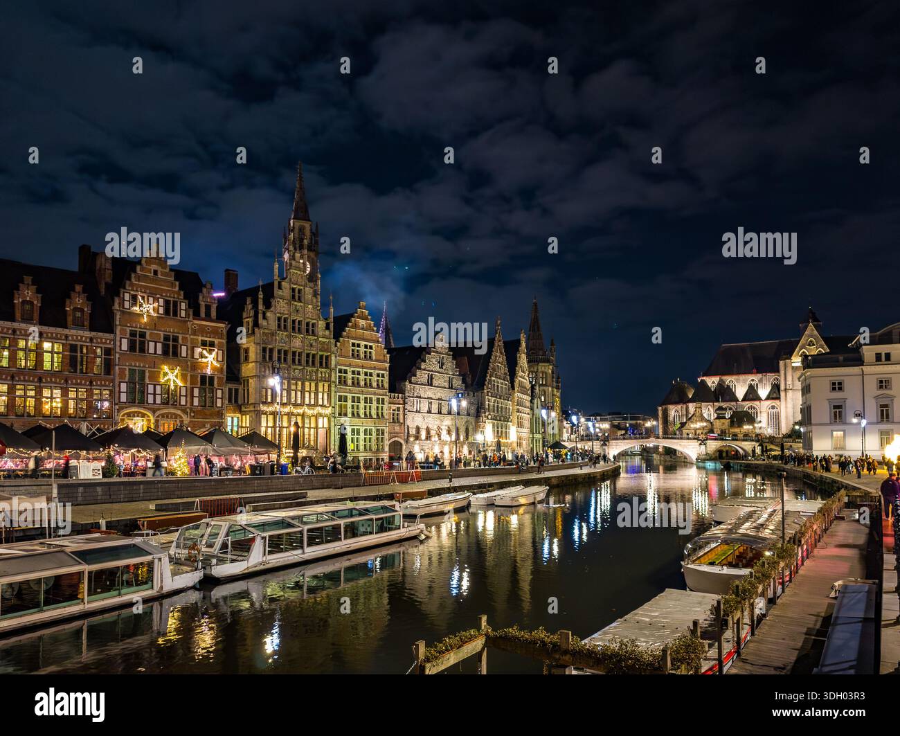 Weihnachtslichter erleuchten die historische Uferpromenade des Flusses Leie in Gent bei Nacht und reflektieren warme, festliche Farben auf dem Wasser, während die Boote durch die fahren Stockfoto