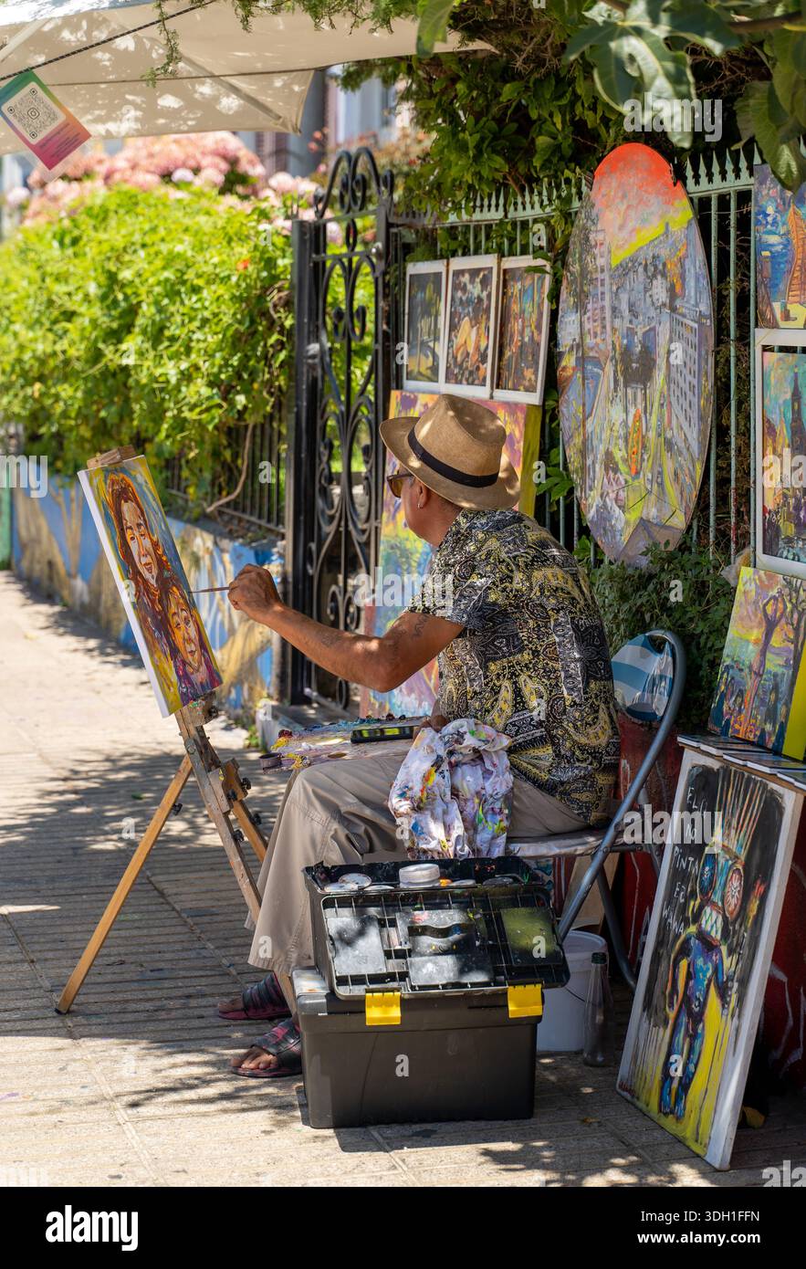 Ein Straßenkünstler malt an einer Staffelei in Valparaíso, umgeben von farbenfrohen Kunstwerken, die entlang eines Bürgersteigs ausgestellt werden. Stockfoto
