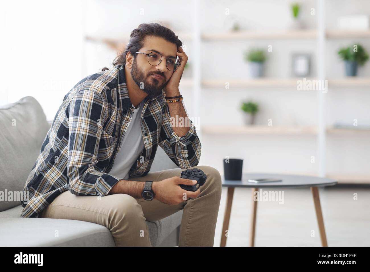 Gelangweilter indischer Kerl, der mit Joystick auf der Couch sitzt Stockfoto