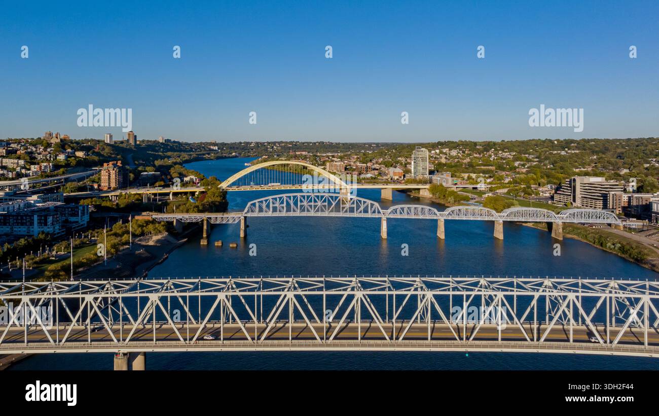 Aus der Vogelperspektive sehen Brücken, die den dunkelblauen Fluss überspannen und den klaren Himmel reflektieren und die Stadtlandschaft mit ihren üppig grünen Ufern verbinden, Cincinnati, Ohio, UN Stockfoto