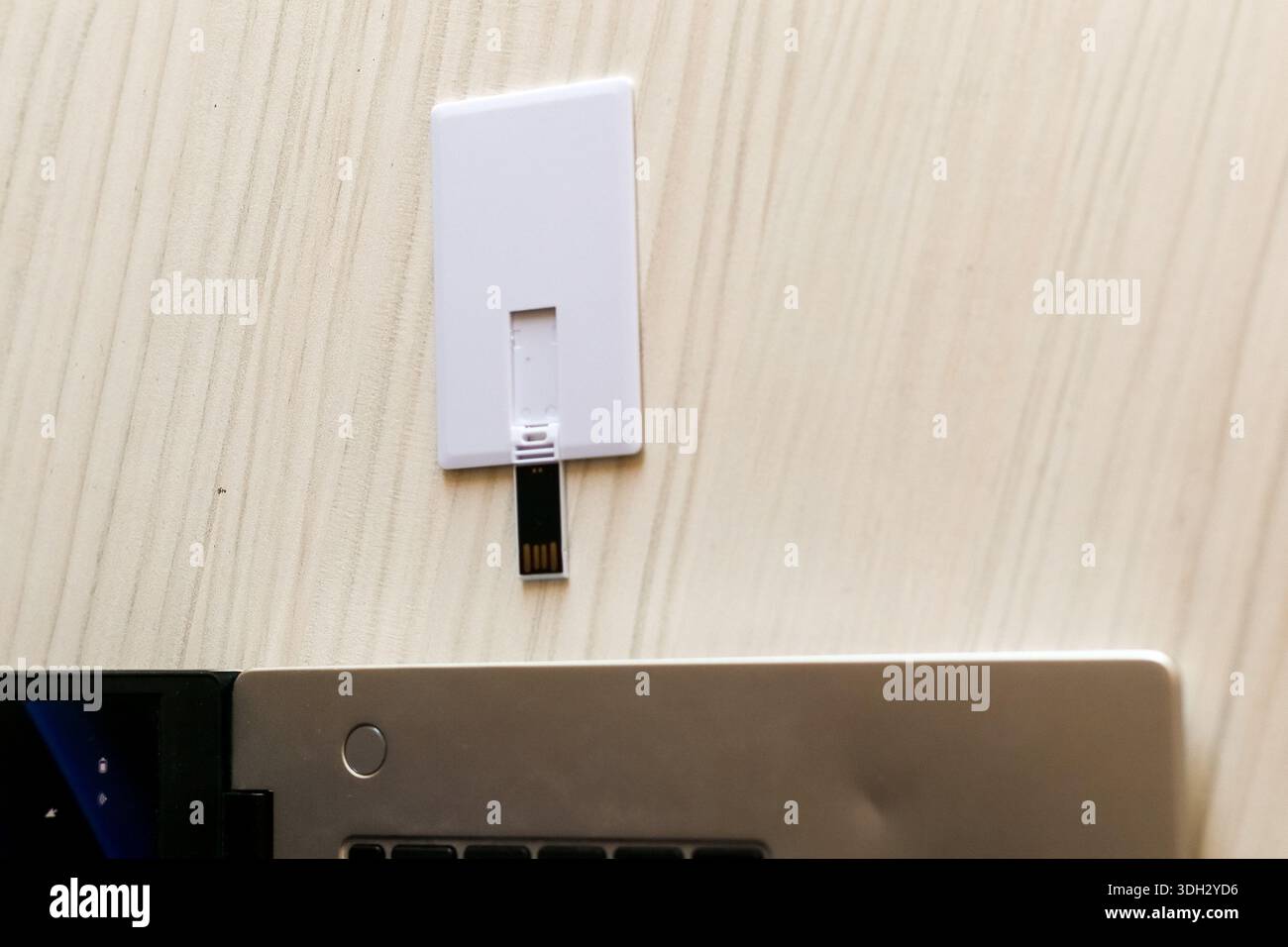 Flaches weißes USB-Kartenlaufwerk mit einem abstehenden schwarzen Anschluss, das sich neben der silbernen Laptop-Tastatur auf einem hellen Holztisch befindet, um schnell zu bedienen. Stockfoto