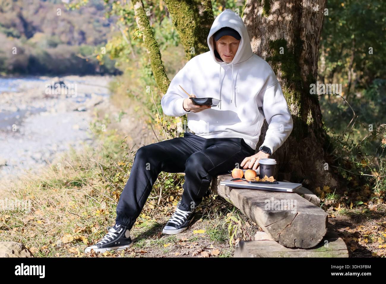 Entspanntes Essen im Freien: Mann auf Holzbank isst Essen, mit Tech-Gadget, Obst, Getränk in Waldumgebung. Stockfoto