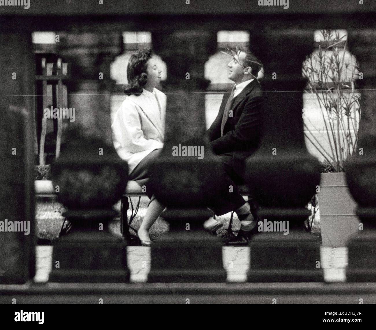 Das Foto zeigt einen Blick durch eine Steinschiene auf einen Mann und eine Frau, die auf einer Bank sitzen und sich zugewandt sind. Rizzuto, Angelo, 1906-1967, Fotograf. 1956. Stockfoto