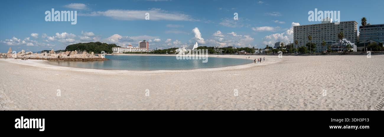 Shirarahama, Japan - 25. Oktober 2023: Panoramablick auf einen breiten weißen Sandstrand, der sich um eine Bucht mit türkisfarbenem Wasser, felsigem Wellenbrecher auf der linken Seite und mehrere moderne Gebäude und Hotels im Hintergrund unter blauem Himmel mit verstreuten Wolken dreht. Stockfoto