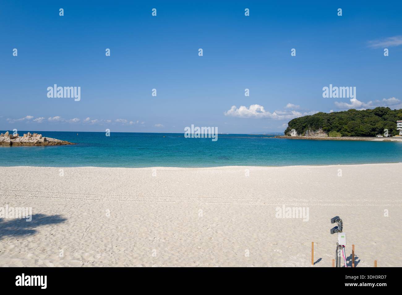 Shirarahama, Japan - 25. Oktober 2023: Ein weiträumiger weißer Sandstrand mit türkisfarbenem Wasser, einem felsigen Wellenbrecher auf der linken Seite und einer bewaldeten Landzunge mit ein paar Gebäuden auf der rechten Seite unter einem klaren blauen Himmel. Stockfoto