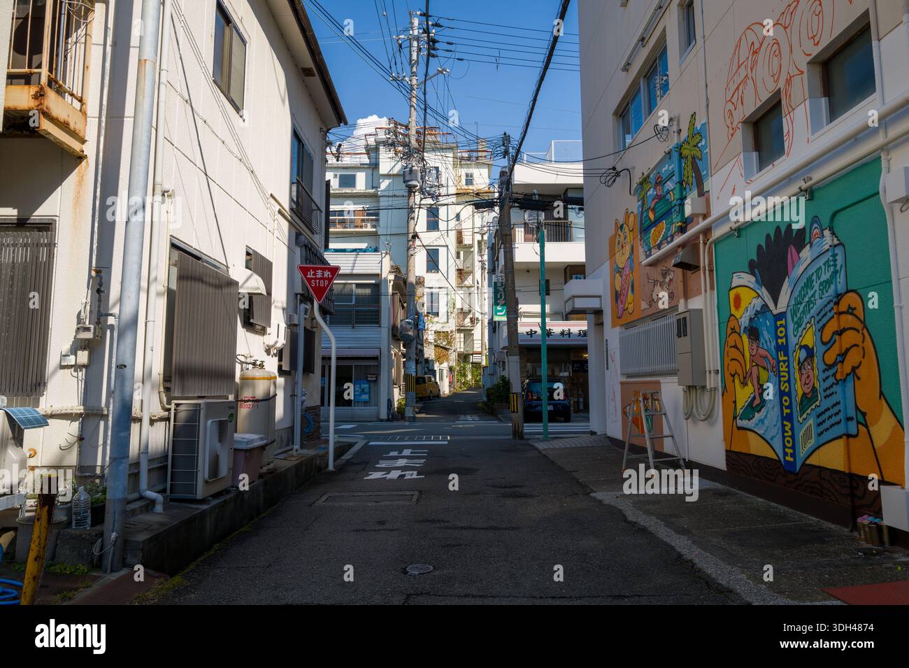 Shirarahama, Japan - 25. Oktober 2023: Eine enge Stadtgasse zeigt bunte Wandmalereien am rechten Gebäude, ein japanisches Stoppschild, Strommasten mit Freileitungen und flache Wohn- und Geschäftsgebäude unter klarem blauen Himmel. Stockfoto