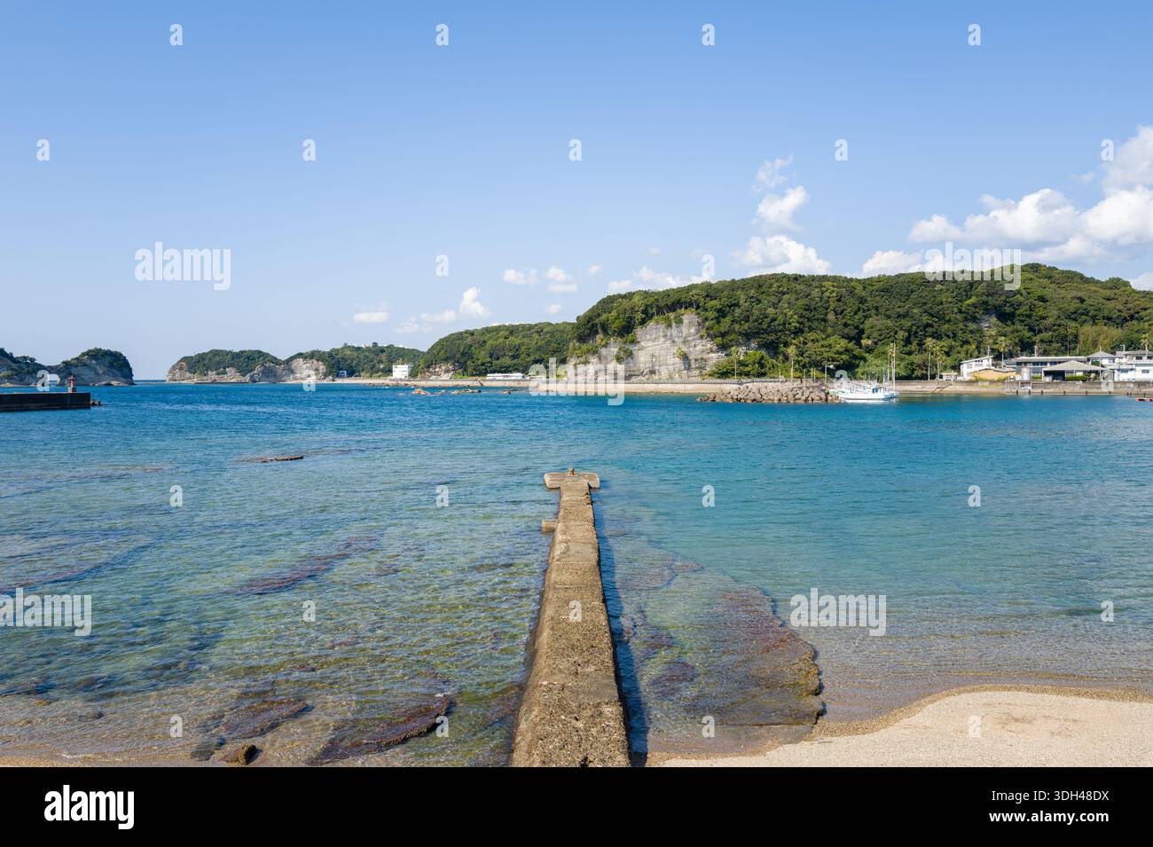 Shirarahama, Japan - 25. Oktober 2023: Ein Betonpier erstreckt sich im Hafen von Shirarahama in klares blaues Wasser, mit bewaldeten Hügeln, felsiger Küste und mehreren Booten und Gebäuden, die im Hintergrund unter klarem Himmel zu sehen sind. Stockfoto