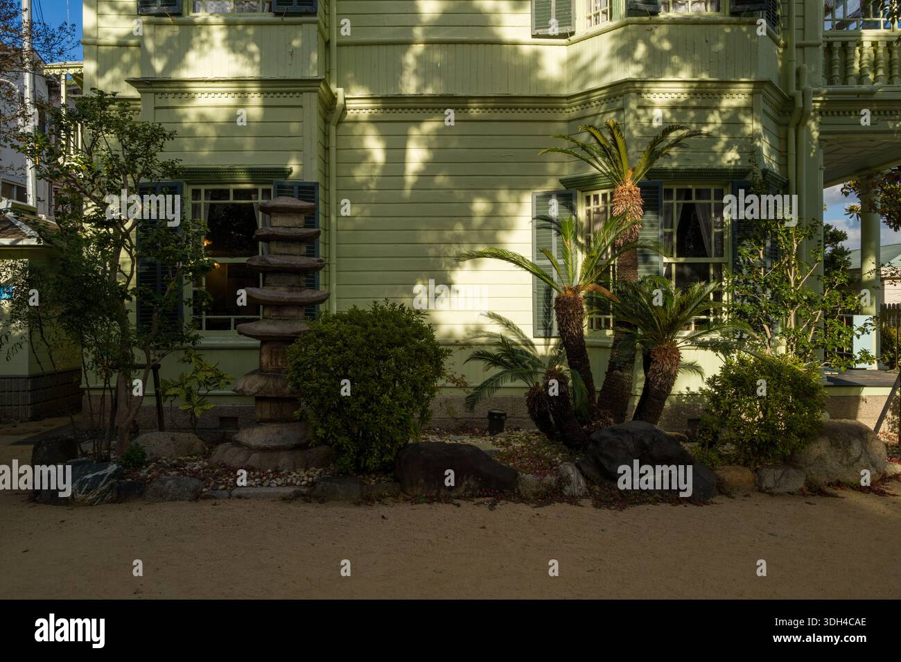 Kobe, Japan - 30. Oktober 2023: Der Garten vor dem Moegi House verfügt über eine mehrstufige Steinpagode, Palmen, Sträucher, Felsen und die hellgrüne Holzfassade mit Fensterläden im Hintergrund. Stockfoto