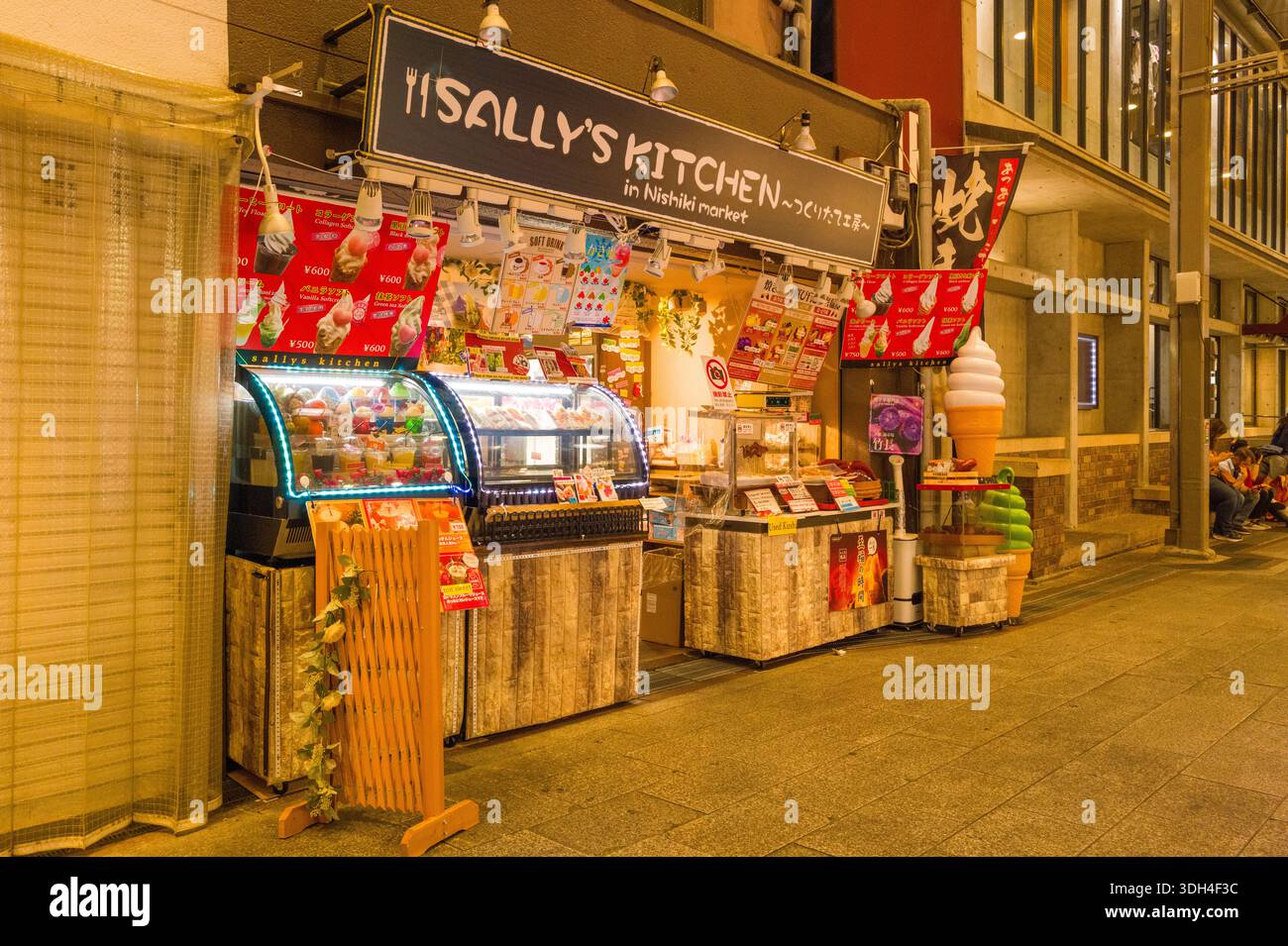 Kyoto, Japan - 19. Oktober 2023: Das Bild zeigt den Essensstand Sally's Kitchen am Nishiki Market in Kyoto bei Nacht mit beleuchteten Vitrinen, farbenfrohen Schildern für Desserts und Eis und einem großen Plastikeiskegel-Modell außerhalb des Ladens. Stockfoto
