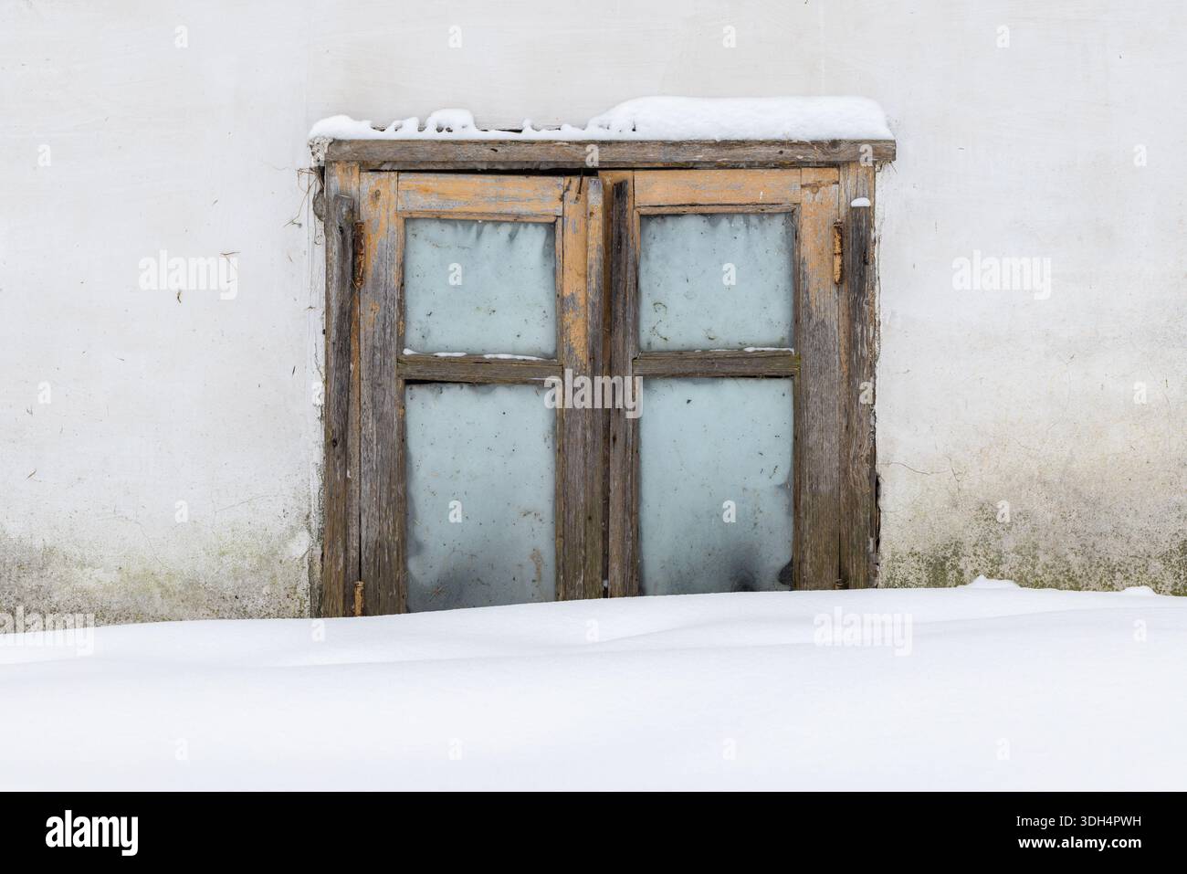 Ein rustikales, verwittertes Holzfenster, eingebettet in eine weiße Wand mit Schnee. Milchglasplatten spiegeln eine ruhige Winterszene wider, die Einsamkeit, Farbe, vermittelt Stockfoto
