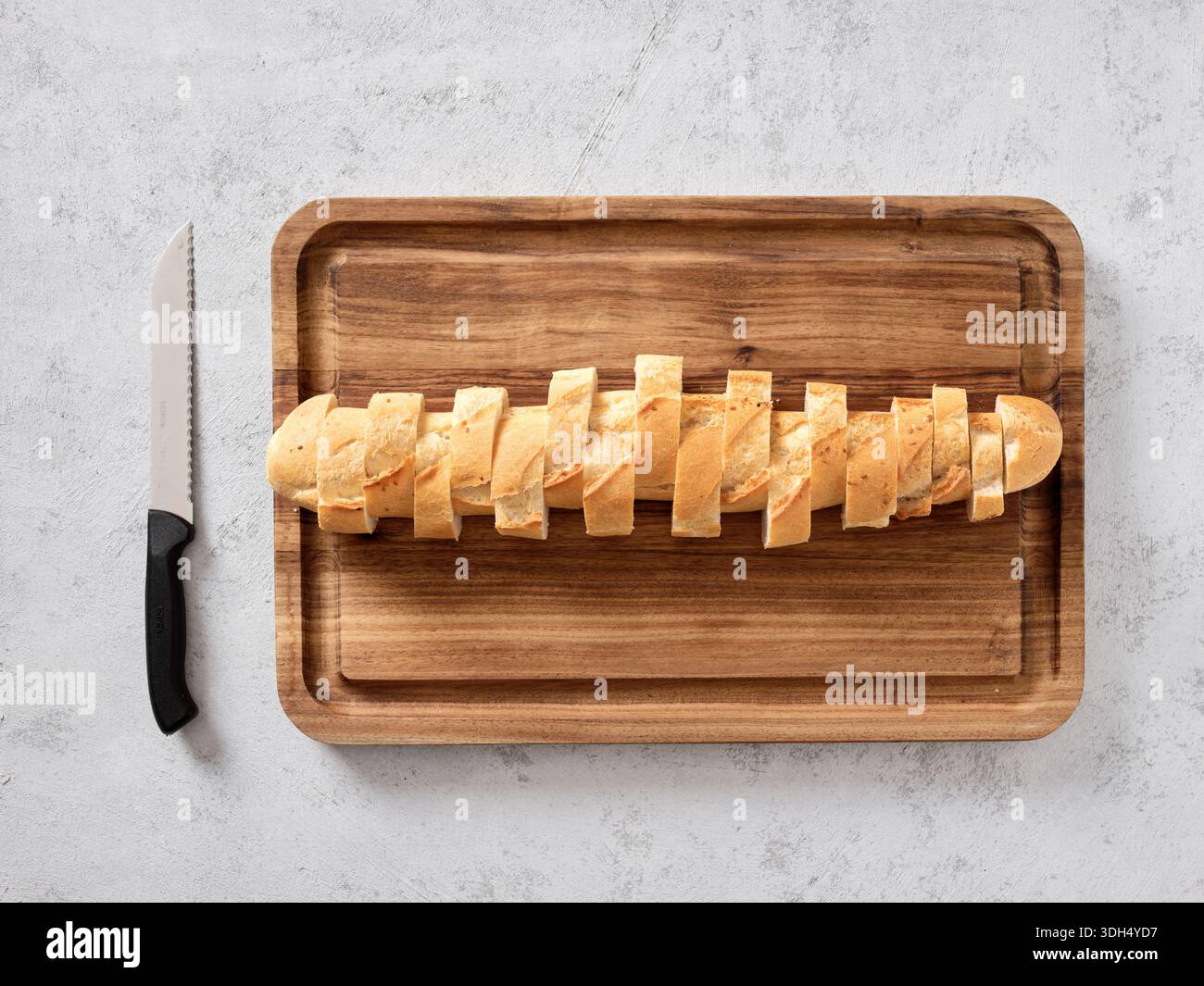 Baguette Brotbrot teilweise auf einem hölzernen Schneidebrett mit einem gezackten Messer geschnitten, von oben gesehen Stockfoto