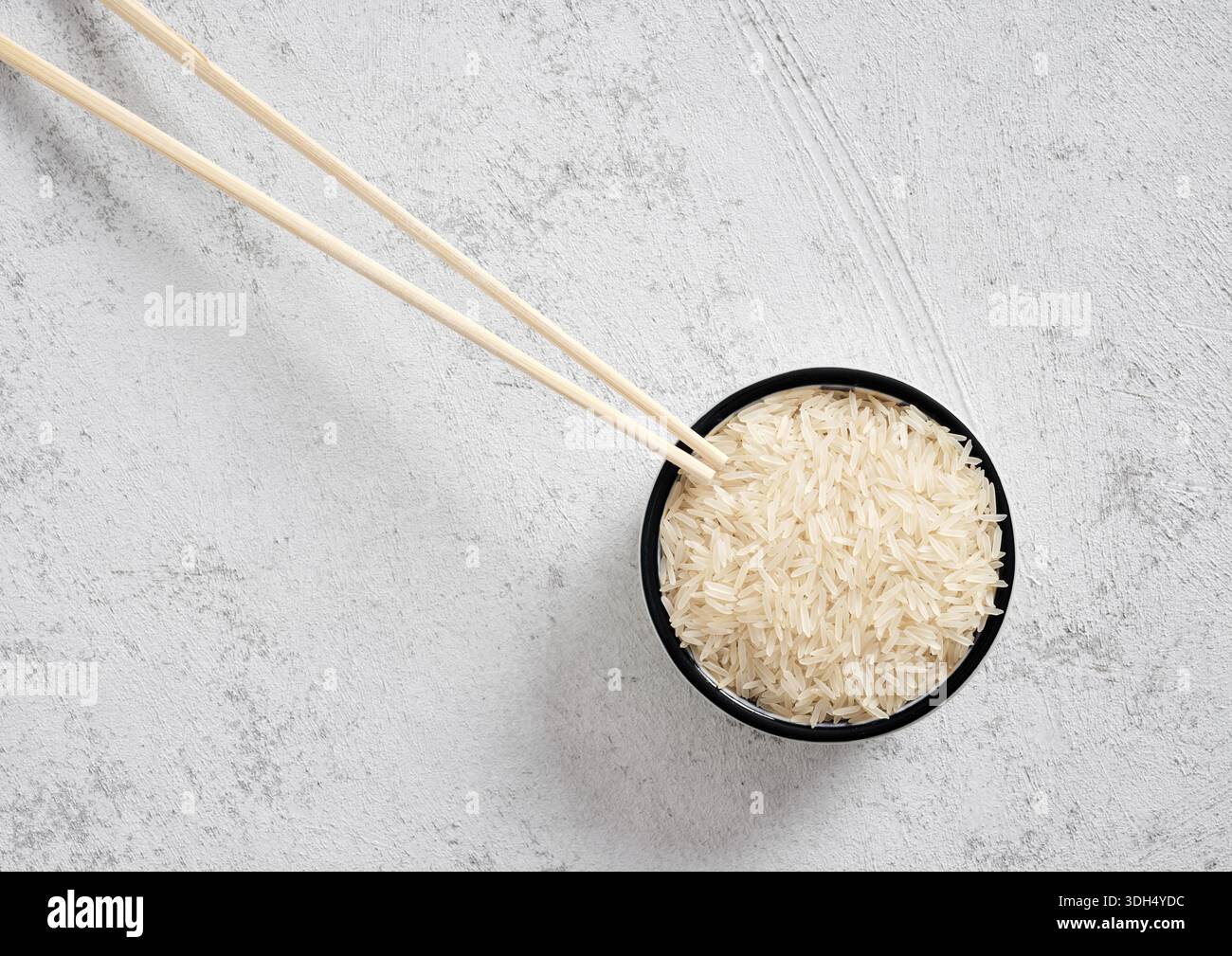 Schüssel mit rohem langkörnigem Basmatireis mit Holzstäbchen auf weißem Betonhintergrund. Flaches, minimales, natürliches Grundnahrungsmittel zum Kochen Stockfoto