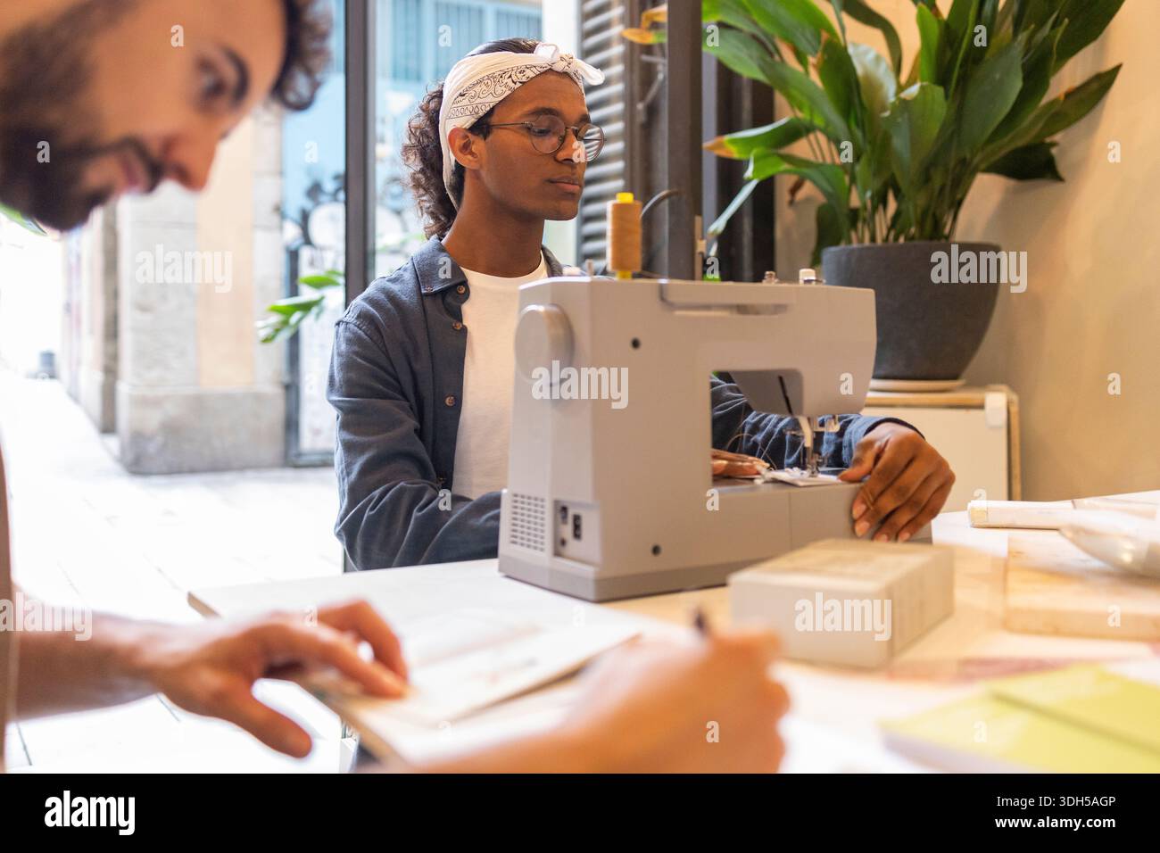 Zwei Kollegen arbeiten in einem stilvollen Coworking-Bereich zusammen. Der eine konzentriert sich auf das Nähen, der andere macht sich Notizen. Natürliches Licht und Pflanzen schaffen eine vibra Stockfoto