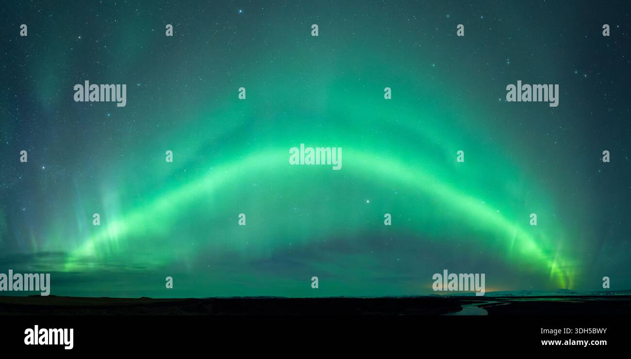 Ein atemberaubender Panoramablick auf die aurora Borealis, der den Nachthimmel mit leuchtenden Grüntönen beleuchtet. Sterne leuchten hell über einem fernen, ruhigen Land Stockfoto