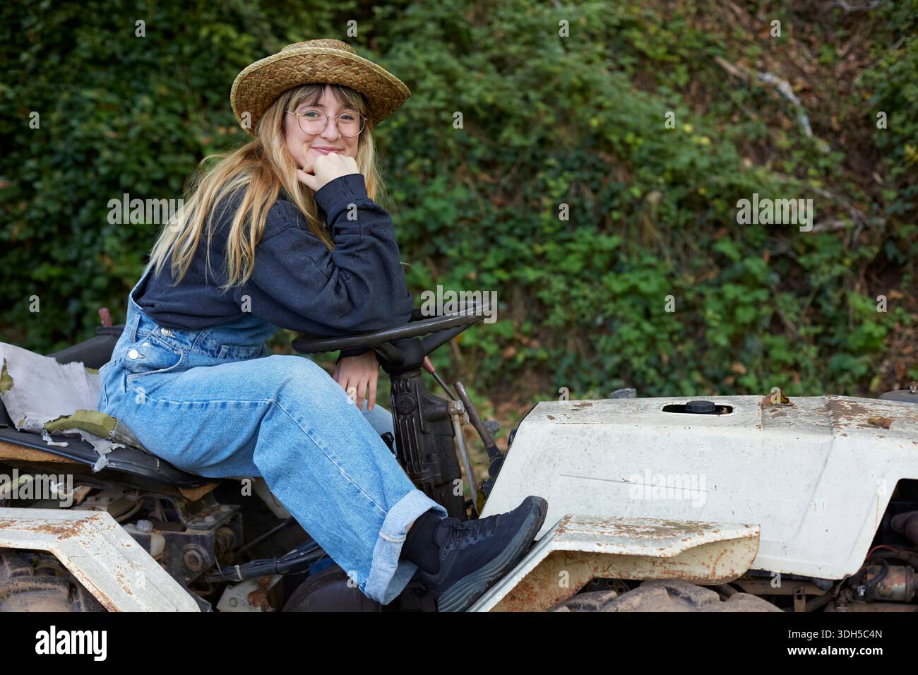 Frau in legeren Kleidern mit Strohhut sitzt auf einem alten weißen Traktor in einer üppigen Gartenumgebung und präsentiert eine Mischung aus Vintage-Maschinen und Natur Stockfoto