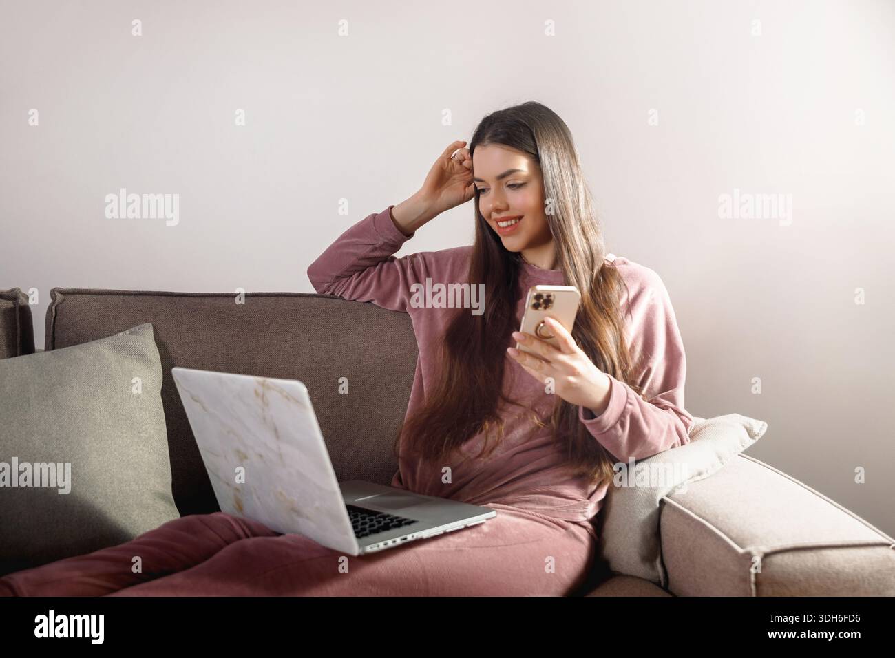 Eine Frau sitzt auf einer Couch und schaut ihr Smartphone mit einem Lächeln an. Ein Laptop ist neben ihr offen. Sonnenlicht erfüllt den Raum, während sie mit ihren Geräten in Berührung kommt Stockfoto