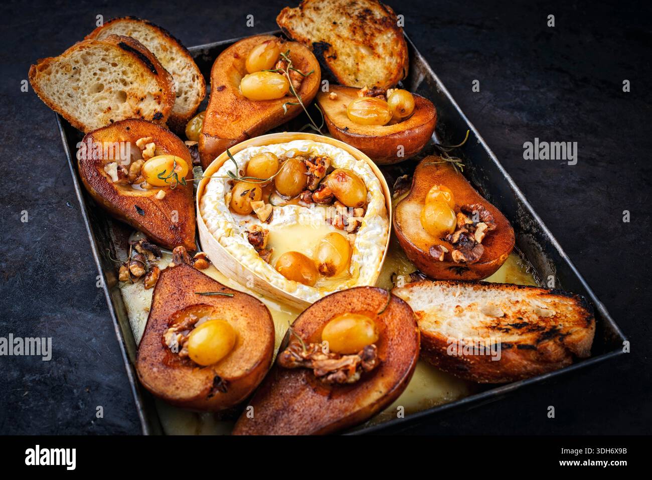 Im Ofen gebackener Camembert-Käse mit karamelisierten glasierten Birnen, Walnüssen und Trauben sowie Baguette auf rustikalem Metallteller Stockfoto