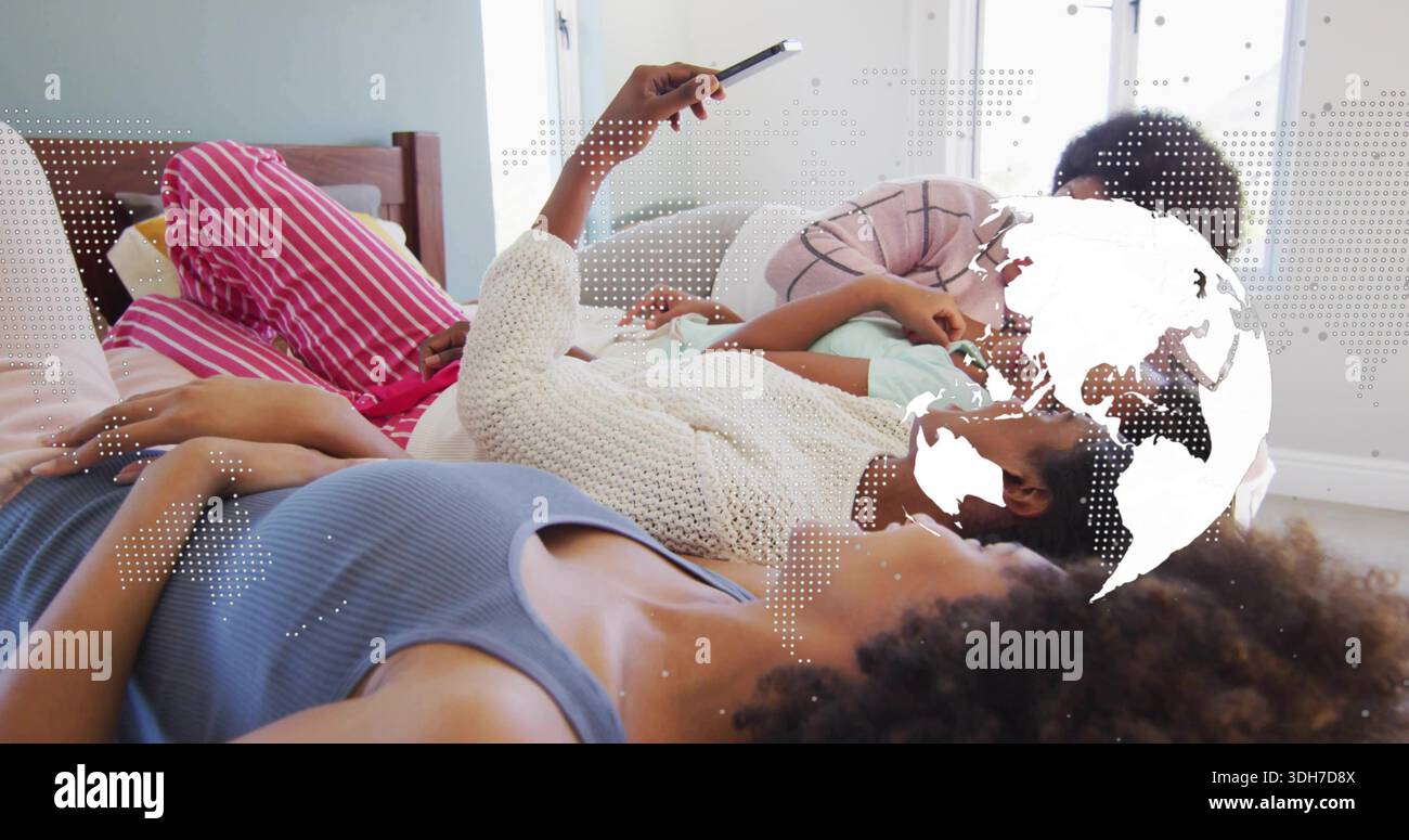 Lachende vier Frauen in Nachtwäsche, die auf dem Bett liegen, das Smartphone hochheben und eine gepunktete Globus-Überlagerung zeigen Stockfoto