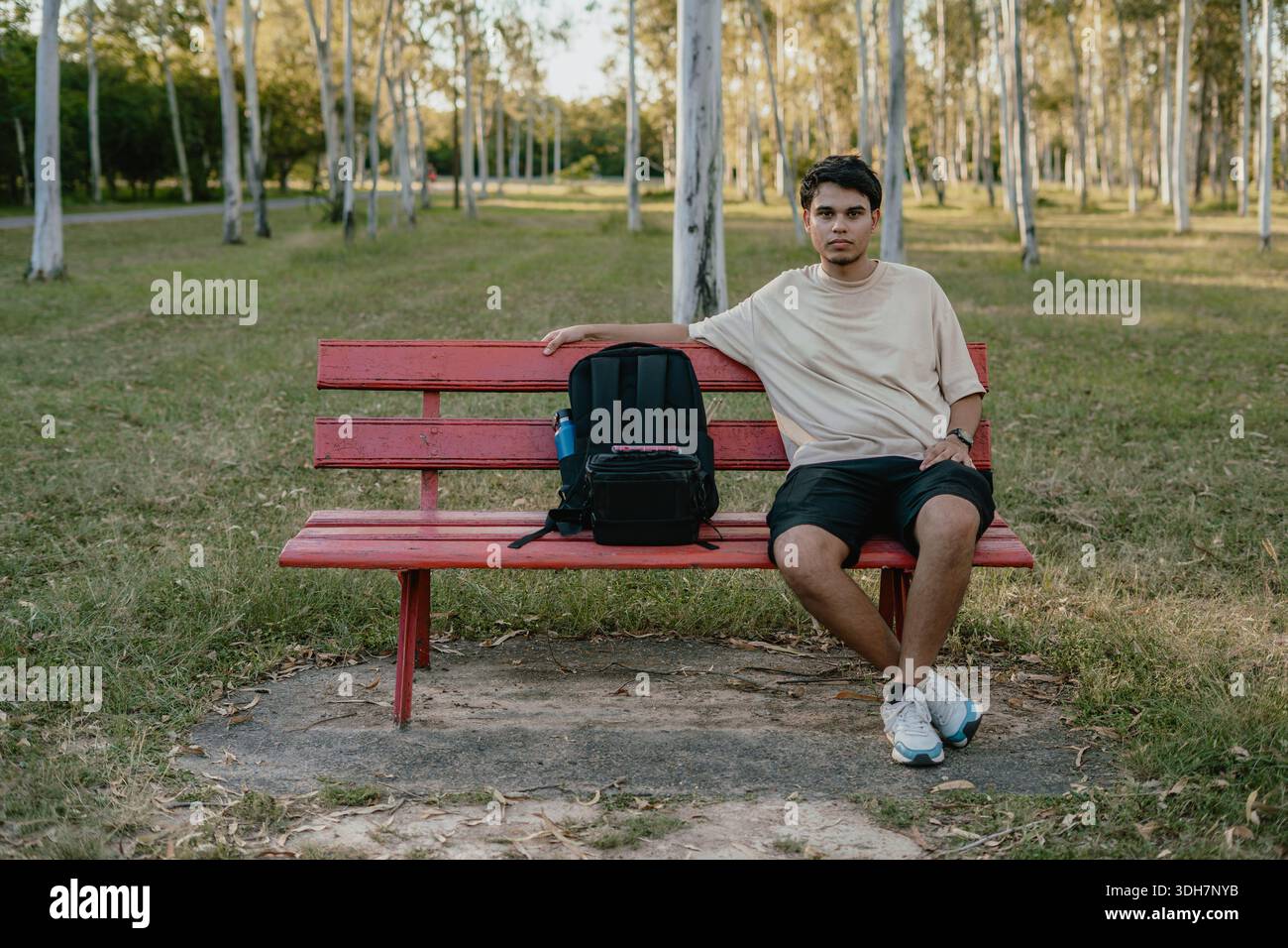 Junger Mann sitzt auf einer roten Bank neben einem Rucksack in einem Park, entspanntes Outdoor-Porträt. Stockfoto