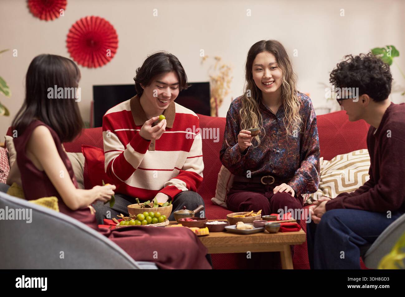Gruppe junger erwachsener asiatischer Männer und Frauen, die zusammen auf dem Sofa sitzen und während der Tet-Feier traditionelle Weihnachtsessen und -Getränke genießen, lächeln und miteinander interagieren Stockfoto