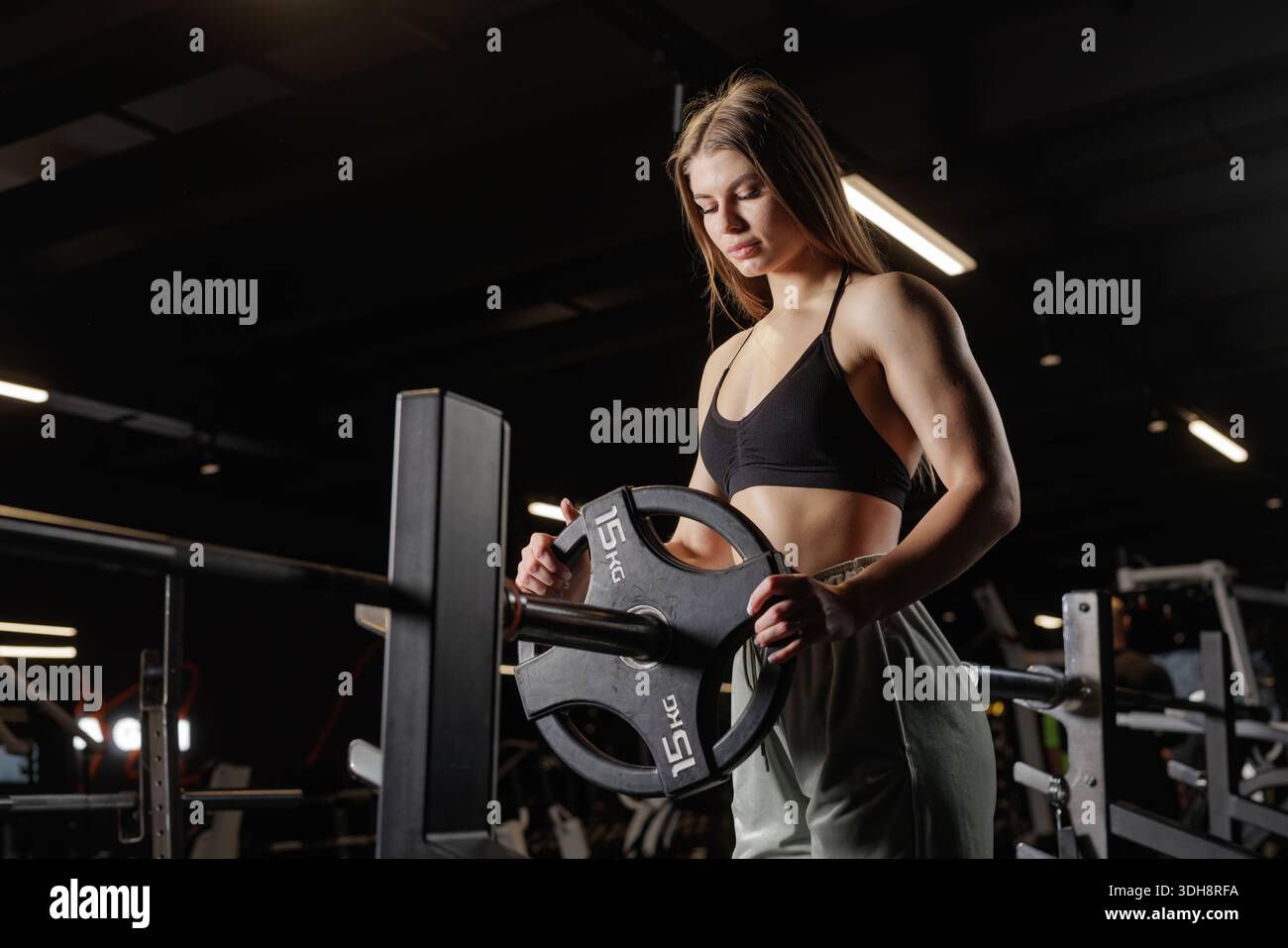 Eine sportliche Frau fügt 15 kg Teller auf die Langhantelablage, um sich für das Krafttraining im Fitnessstudio vorzubereiten Stockfoto