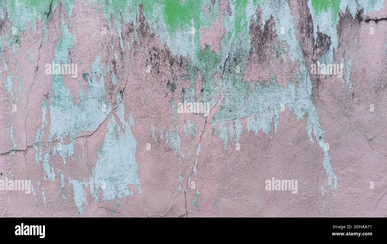 Eine verwitterte Betonwand zeigt strukturierte Schichten von abblätternder rosa und grüner Farbe, die Risse, Schimmel und Anzeichen von Verfall durch Alterung sichtbar machen. Stockfoto