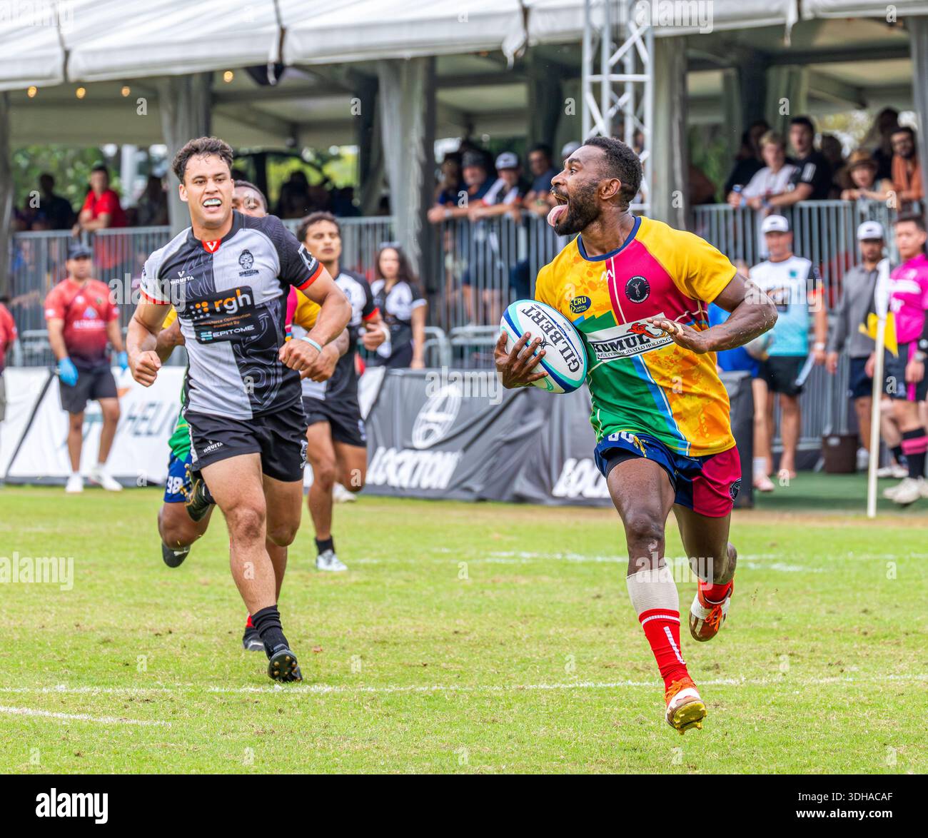 Dynamische Rugby-Siebenkampf, während ein farbenfroher Angreifer während eines Outdoor-Turniers mit dem Ball über die Touchline sprintet, verfolgt von den Verteidigern. Stockfoto