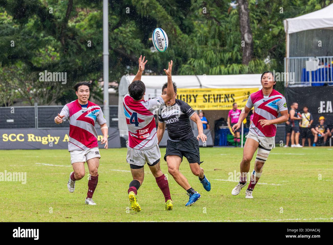 Rugby Sevens Spieler springen, um einen hohen Ball im Mittelfeld während eines Wettkampfes zu messen. Ein Team in rot-weißen Trikots fordert einen schwarzen Shir heraus Stockfoto