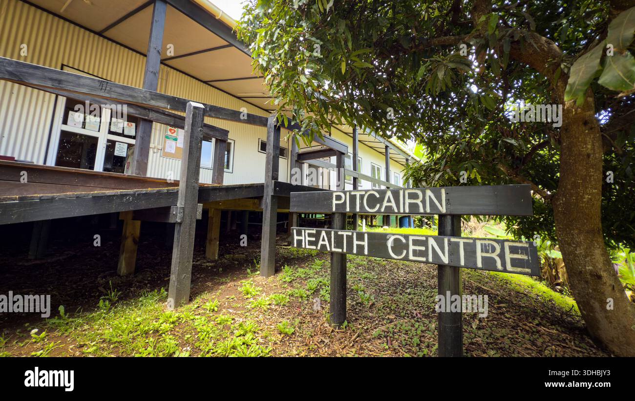 Blick auf das Schild des Pitcairn Health Centre vor einem langen, niedrigen Holzgebäude mit dunklem Dach, eingebettet in üppiges Grün, Adamstown, Pitca Stockfoto