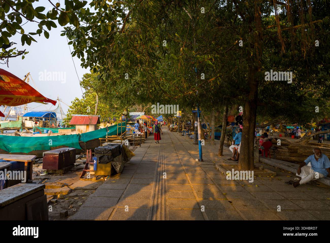Fort Kochi, Indien - 16. März 2023: Mehrere Menschen sitzen und gehen auf einem gepflasterten Pfad, der von großen Bäumen beschattet wird, mit Fischerbooten, Marktständen und verschiedenen Waren, die auf beiden Seiten des Gehwegs zu sehen sind. Stockfoto