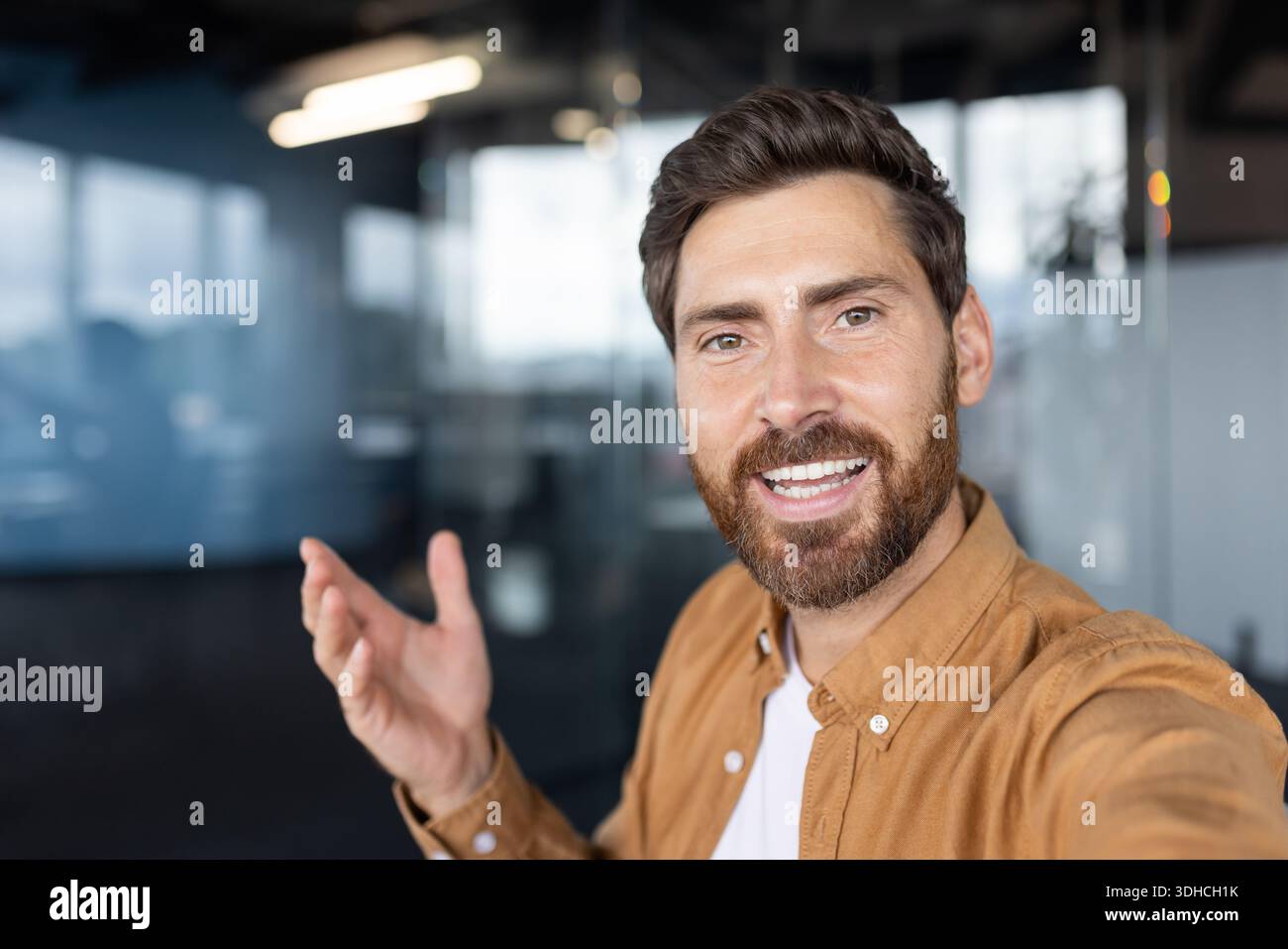 Ein lächelnder bärtiger Mann, der eine einladende Handbewegung macht, während er die Kamera aus der Perspektive der ersten Person betrachtet und das Vertrauen in eine moderne Büroumgebung zum Ausdruck bringt Stockfoto