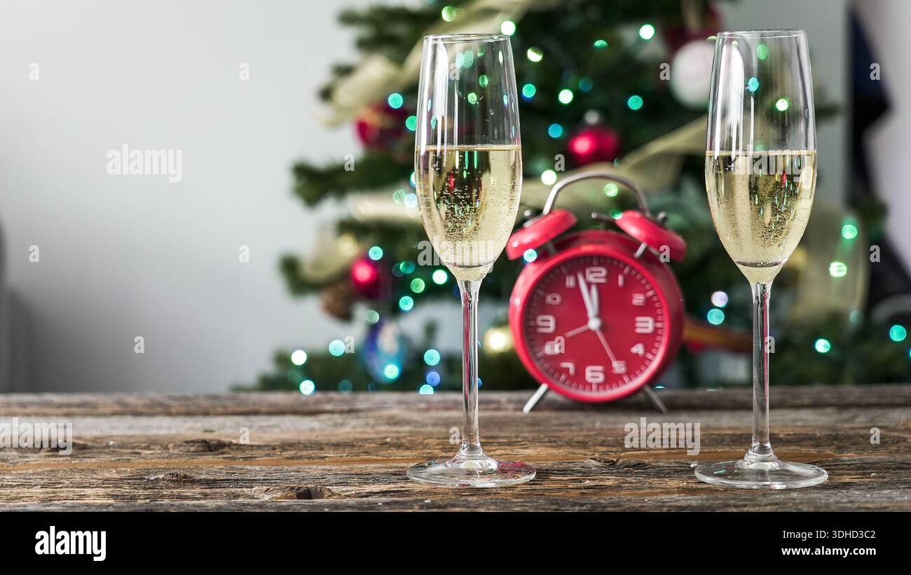 Zwei Sektgläser sitzen auf einem Holztisch. In der Nähe befindet sich eine rote Uhr, die eine festliche Kulisse mit einem mit Lichtern und Ornamenten verzierten Weihnachtsbaum zeigt Stockfoto