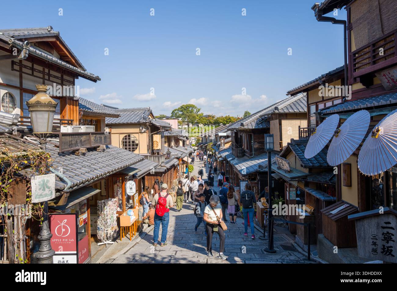 Kyoto, Japan - 16. Oktober 2023: Menschen werden auf den Steintreppen der Ninenzaka-Straße, umgeben von traditionellen Holzhäusern, Ladenschildern und dekorativen Regenschirmen, unter einem klaren blauen Himmel gesehen. Stockfoto