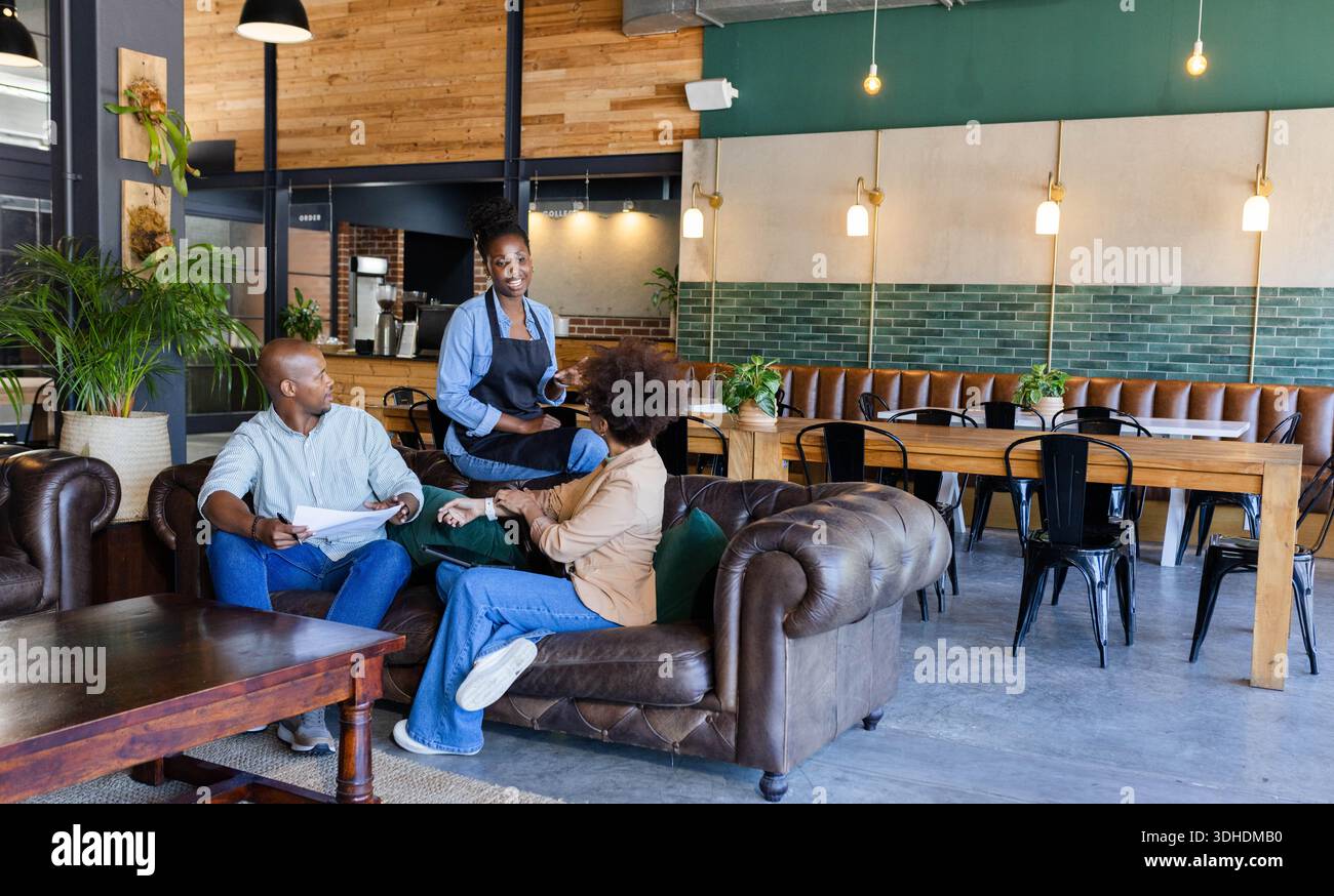 Afroamerikanischer Kellner auf Schürze, Gäste unterhalten sich, halten Menü und Tablett auf dem Sofa im Café Stockfoto