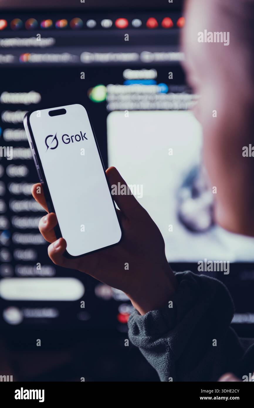 Izmir, Türkei - 21. Januar 2026: Foto einer Frau, die ein modernes Smartphone hält und das Grok-Logo scharf im Fokus zeigt, ein großer Computerbildschirm hinter X Stockfoto