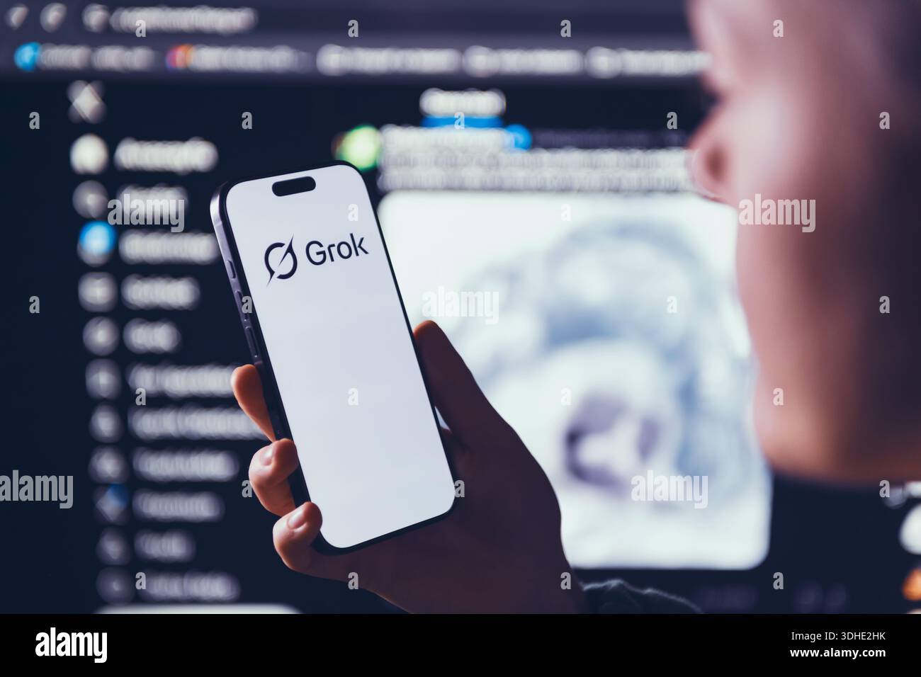 Izmir, Türkei - 21. Januar 2026: Foto einer Frau, die ein modernes Smartphone hält und das Grok-Logo scharf im Fokus zeigt, ein großer Computerbildschirm hinter X Stockfoto