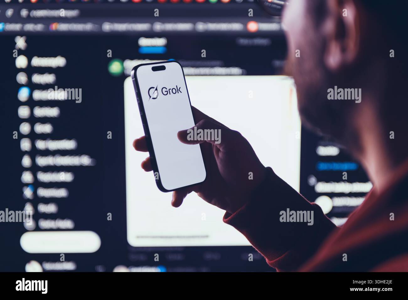 Izmir, Türkei - 21. Januar 2026: Foto eines Mannes, der ein modernes Smartphone hält und das Grok-Logo scharf zeigt, großer Computerbildschirm im Hintergrund Stockfoto
