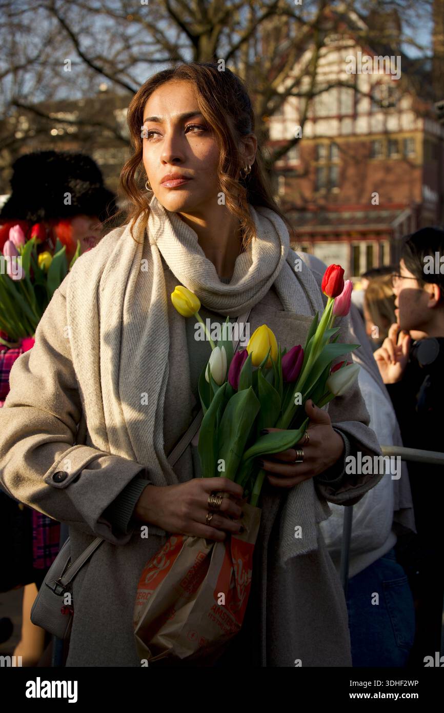 Eine nachdenkliche Frau mit welligen dunklen Haaren hält einen mehrfarbigen Tulpenstrauß beim jährlichen Tulpentag in Amsterdam inmitten einer vielfältigen Menschenmenge und Giebelhäusern Stockfoto