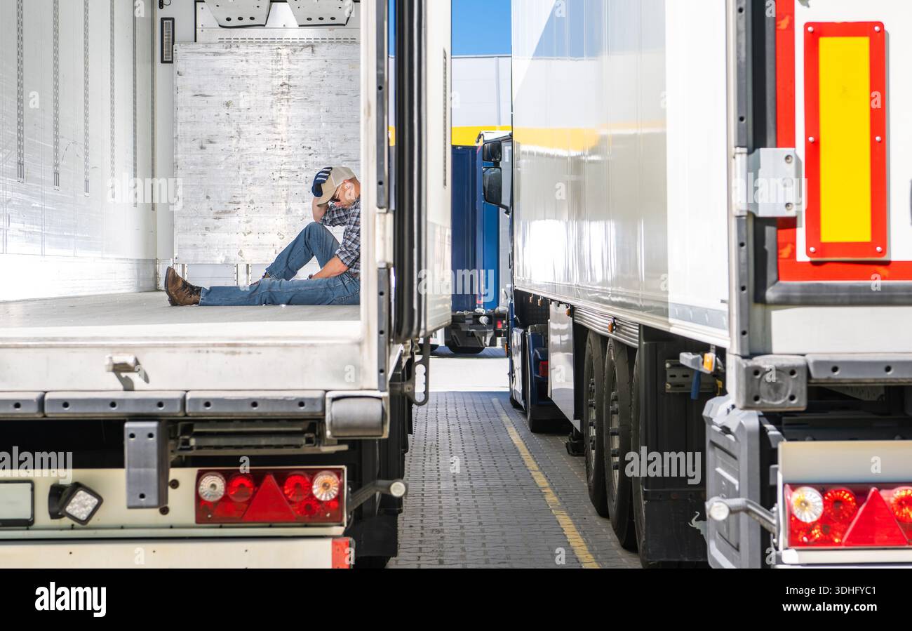 Müder Trcuker sitzt tagsüber auf dem Boden eines Lkw-Anhängers, während er in der Nähe anderer Lkw in einem Laderaum parkt. Stockfoto