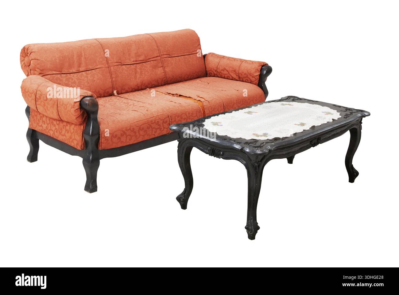 Ein altes orangefarbenes gepolstertes Sofa mit sichtbarer Abnutzung und zerrissenem Stoff steht neben einem klassischen schwarzen Holztisch mit weißer Tischdecke aus Spitze Stockfoto