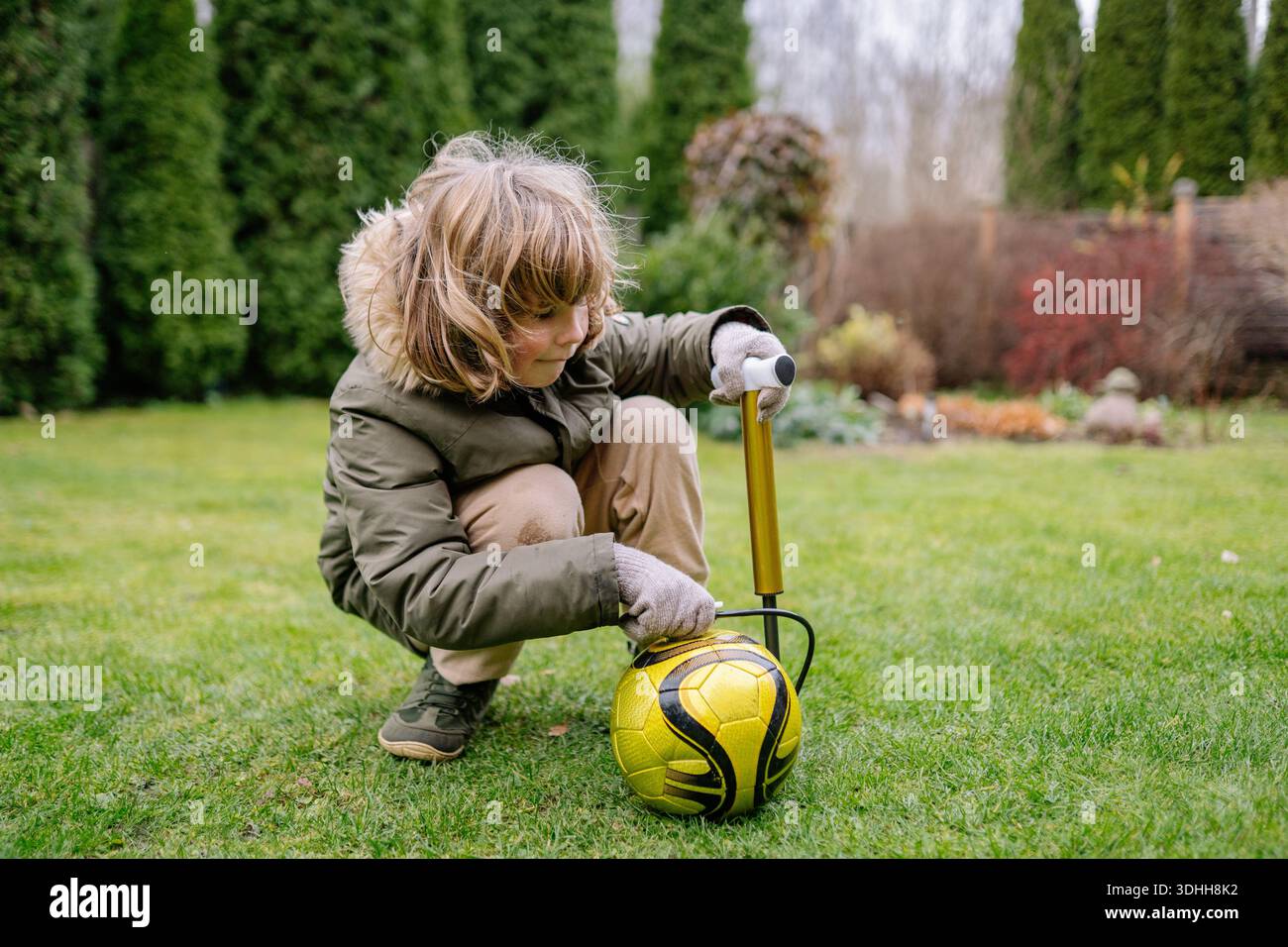 Ein Kind kniet auf Gras im Garten. Das Kind pumpt einen gelben Fußball mit einer Pumpe auf. Bäume und Büsche umgeben die Gegend. Stockfoto